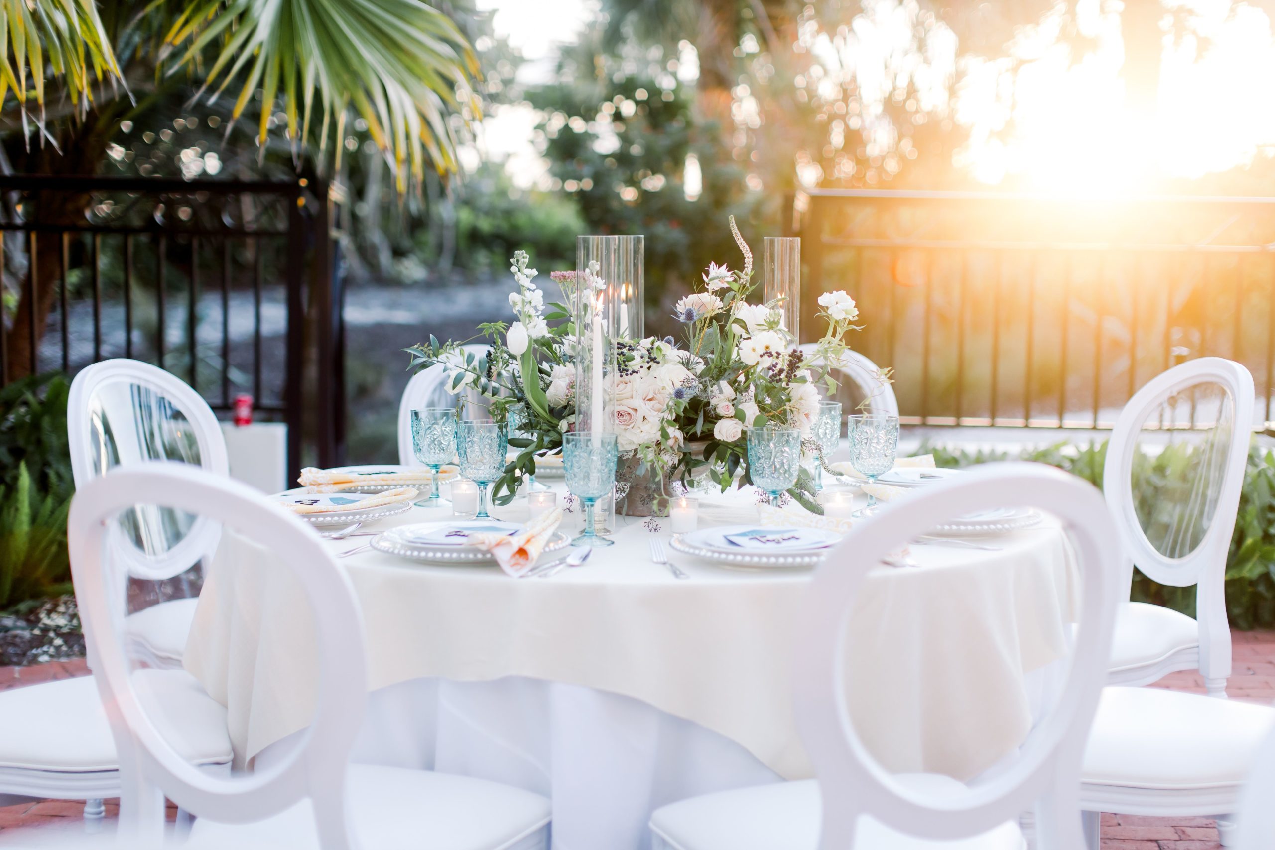 Wedding Dining at Sunset with white chairs and white linens featuring glassware and plates near palm trees at the Sea Palms Estate in Captiva Island, FL