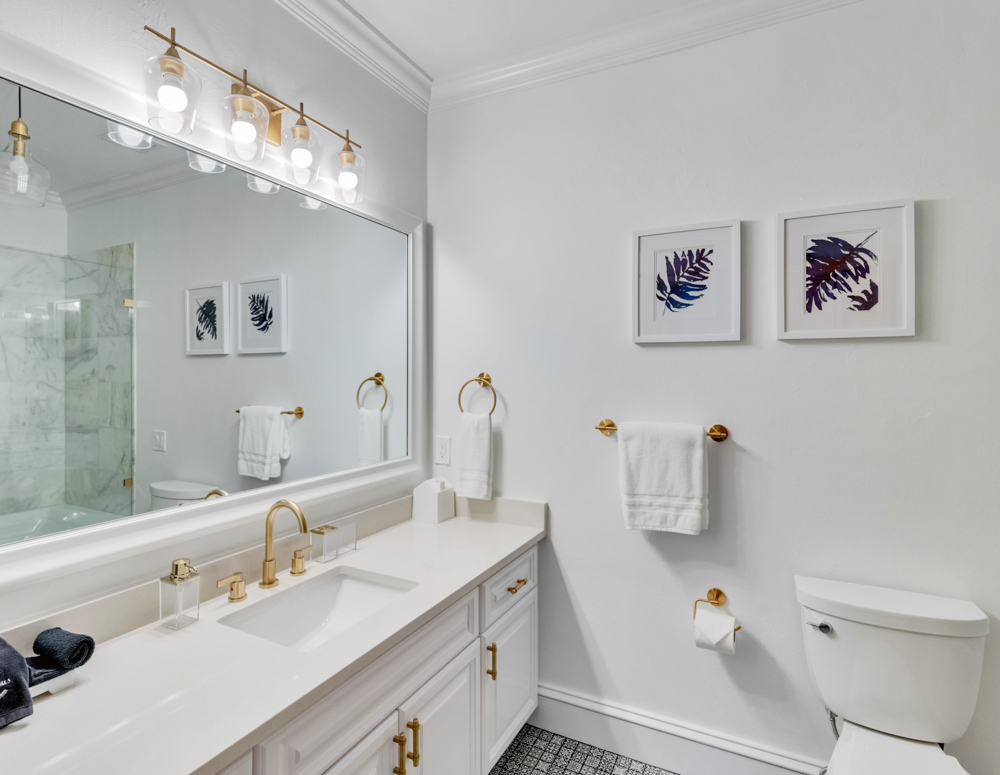 White Bathroom with golden fixtures, white towels and a large mirror along with flower paintings on the wall at the Sea Palms Estate in Captiva Island, FL