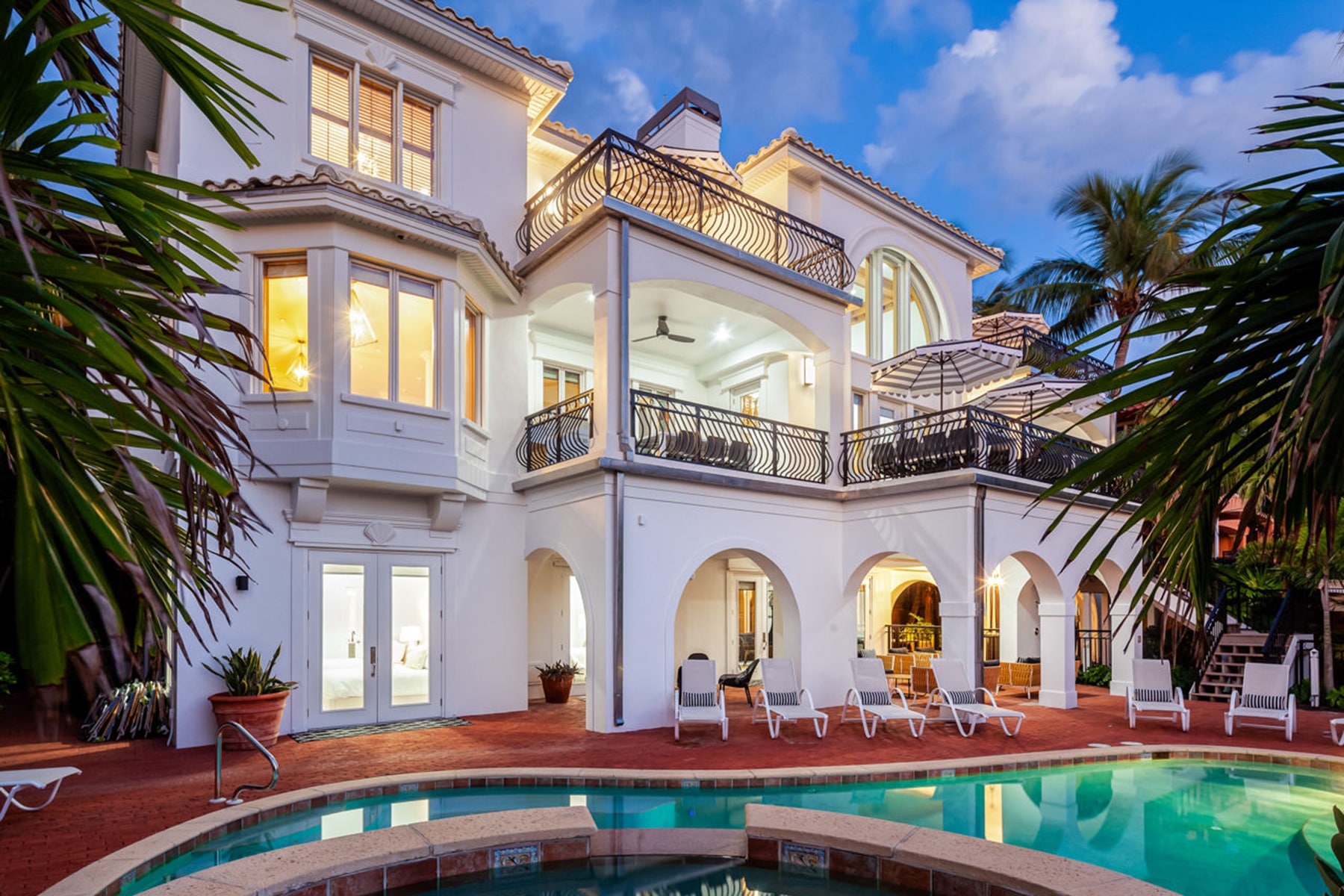A white three floor estate with balconies, patio furniture and blue and white umbrellas, a patio area with a pool, hot tub and pool chairs, and a sandy walk way to the beach surrounded with palm trees in Captiva Island, FL